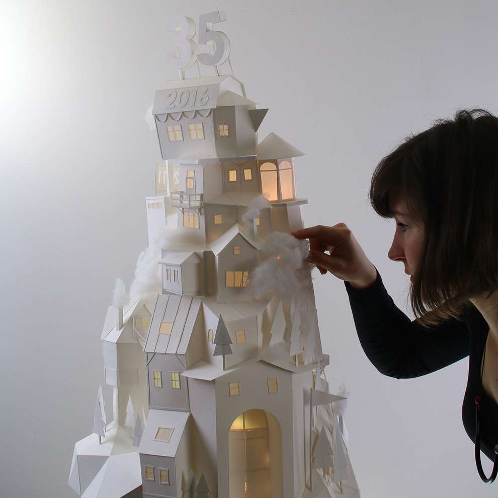 Vera Van Wolferen, Behind the scenes photo of Vera crafting an intricate paper sculpture of a cityscape. Magical miniature paper universe.  